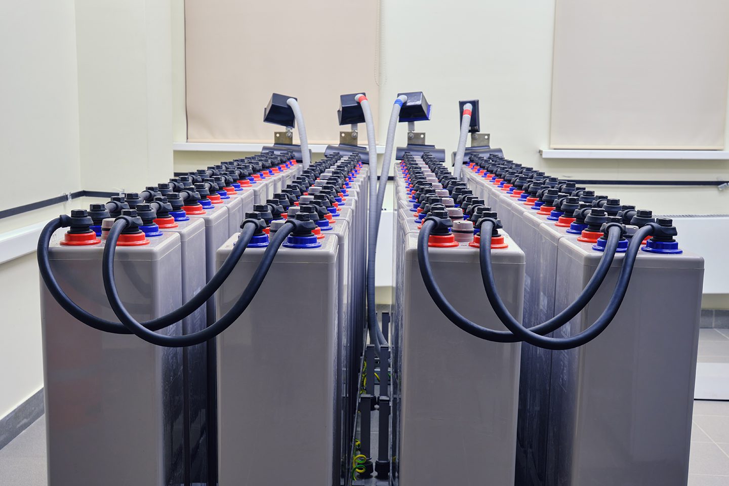 Two rows of industrial batteries, each with attached cables and colored caps, are arranged in a brightly lit room.
