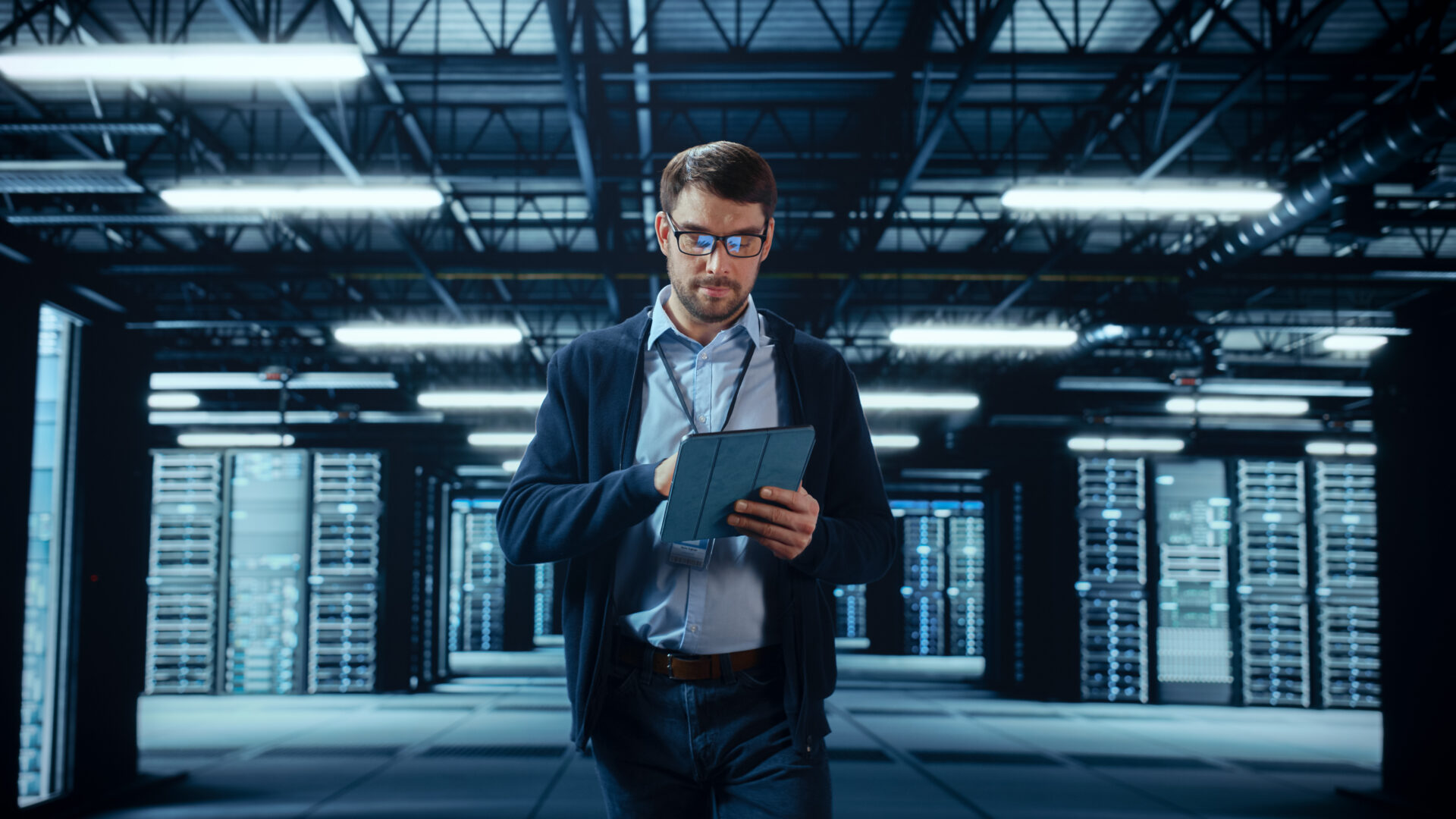 Male IT Specialist Walks Between Row of Operational Server Racks in Data Center. Engineer Uses Tablet Computer for Maintenance. Concept for Cloud Computing, Artificial Intelligence, Cybersecurity. Man in a data center holding a tablet, surrounded by rows of server racks.