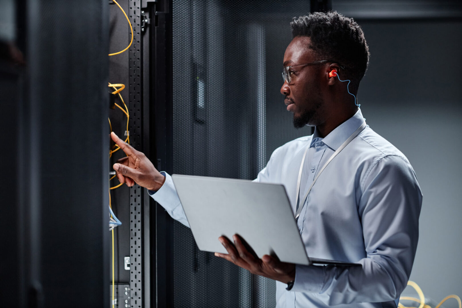Young network engineer working with servers in data center and holding laptop A person wearing glasses and holding a laptop works with cables in a server room, wearing an earbud for communication.