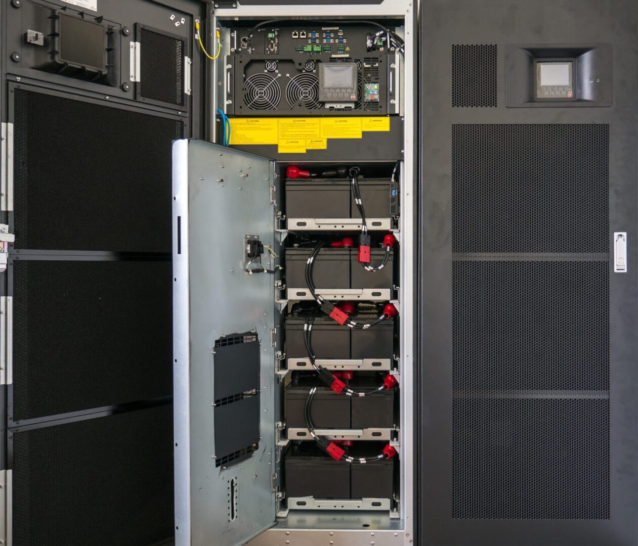 Battery of large uninterruptible Power Supply (UPS) An open server rack displaying multiple black power modules and cables, with a control panel above and ventilation panels on the side.