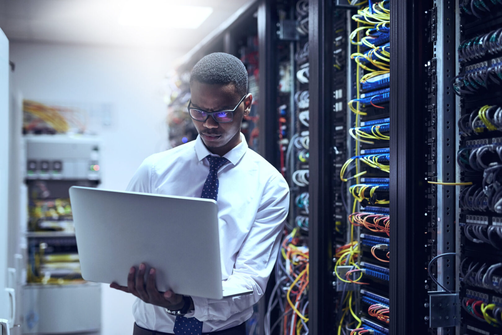 The man with the plan A person in a shirt and tie holds a laptop while standing in a server room surrounded by server racks and cables.